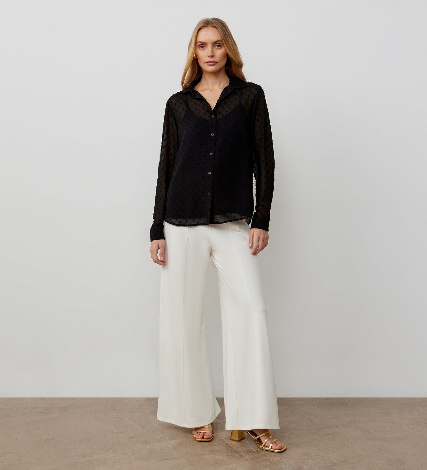 A woman stands against a plain background wearing the Janette Black Chiffon Blouse With Detachable Cami— a sheer black button-up with textured spot detail—paired with white wide-leg pants and gold heeled sandals.