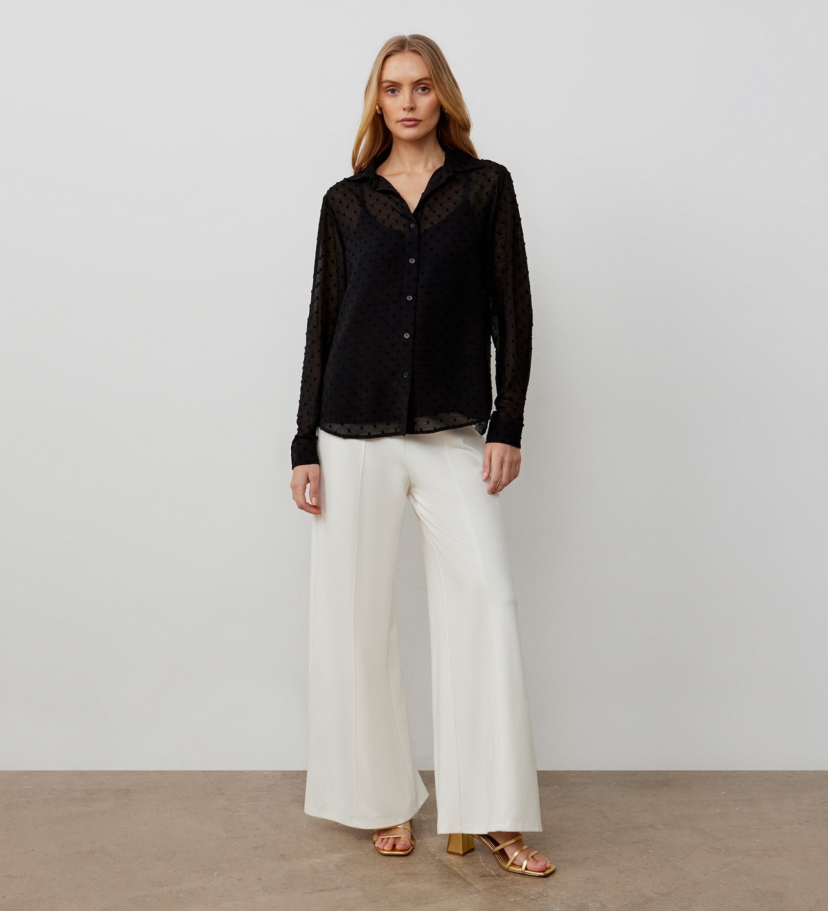 A woman stands against a plain background wearing the Janette Black Chiffon Blouse With Detachable Cami— a sheer black button-up with textured spot detail—paired with white wide-leg pants and gold heeled sandals.