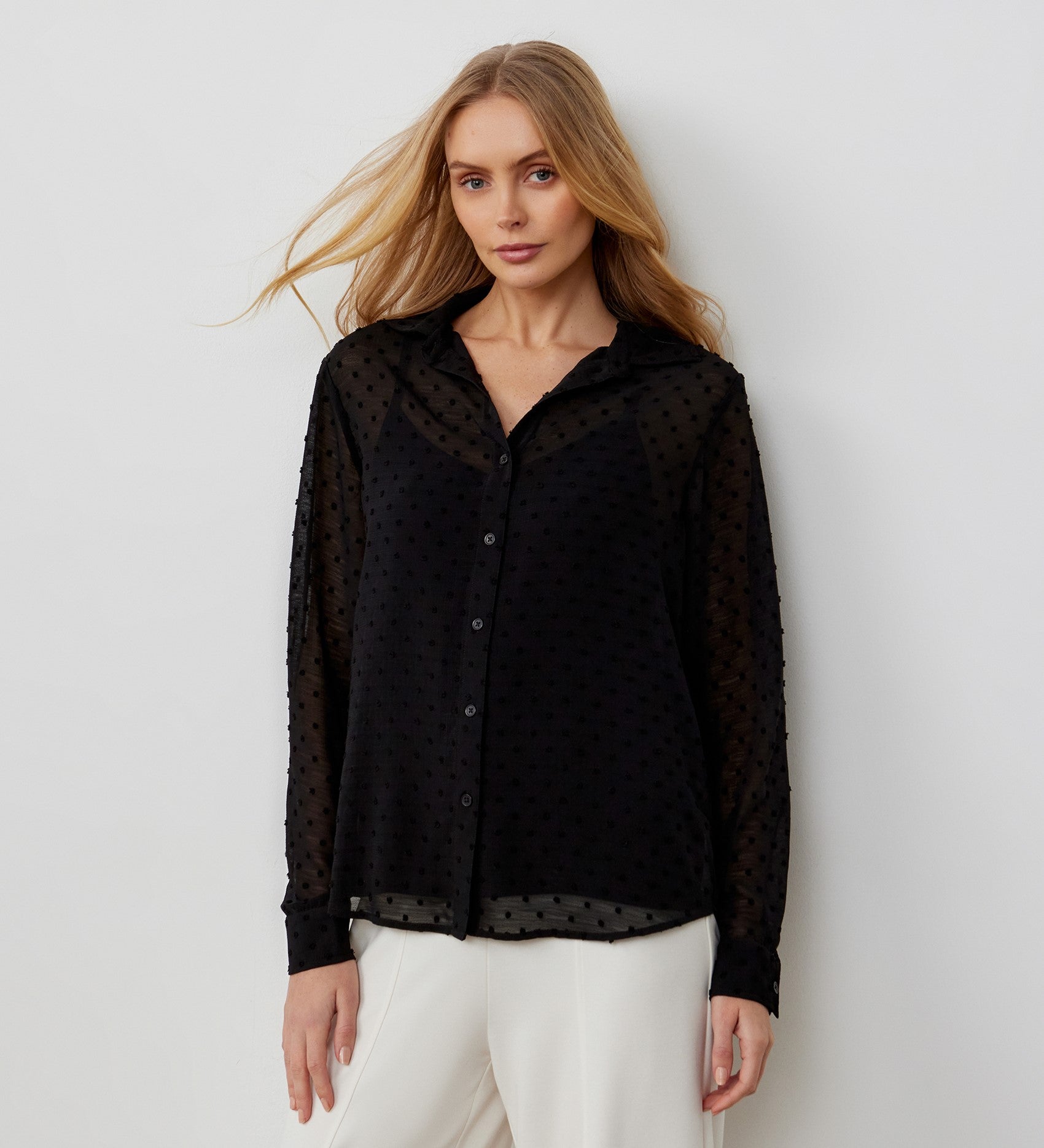 A woman with long blonde hair wears the Janette Black Chiffon Blouse With Detachable Cami, featuring a sheer, textured design and subtle dotted pattern, paired with white pants against a plain light background.