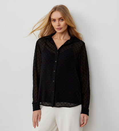 A woman with long blonde hair wears the Janette Black Chiffon Blouse With Detachable Cami, featuring a sheer, textured design and subtle dotted pattern, paired with white pants against a plain light background.