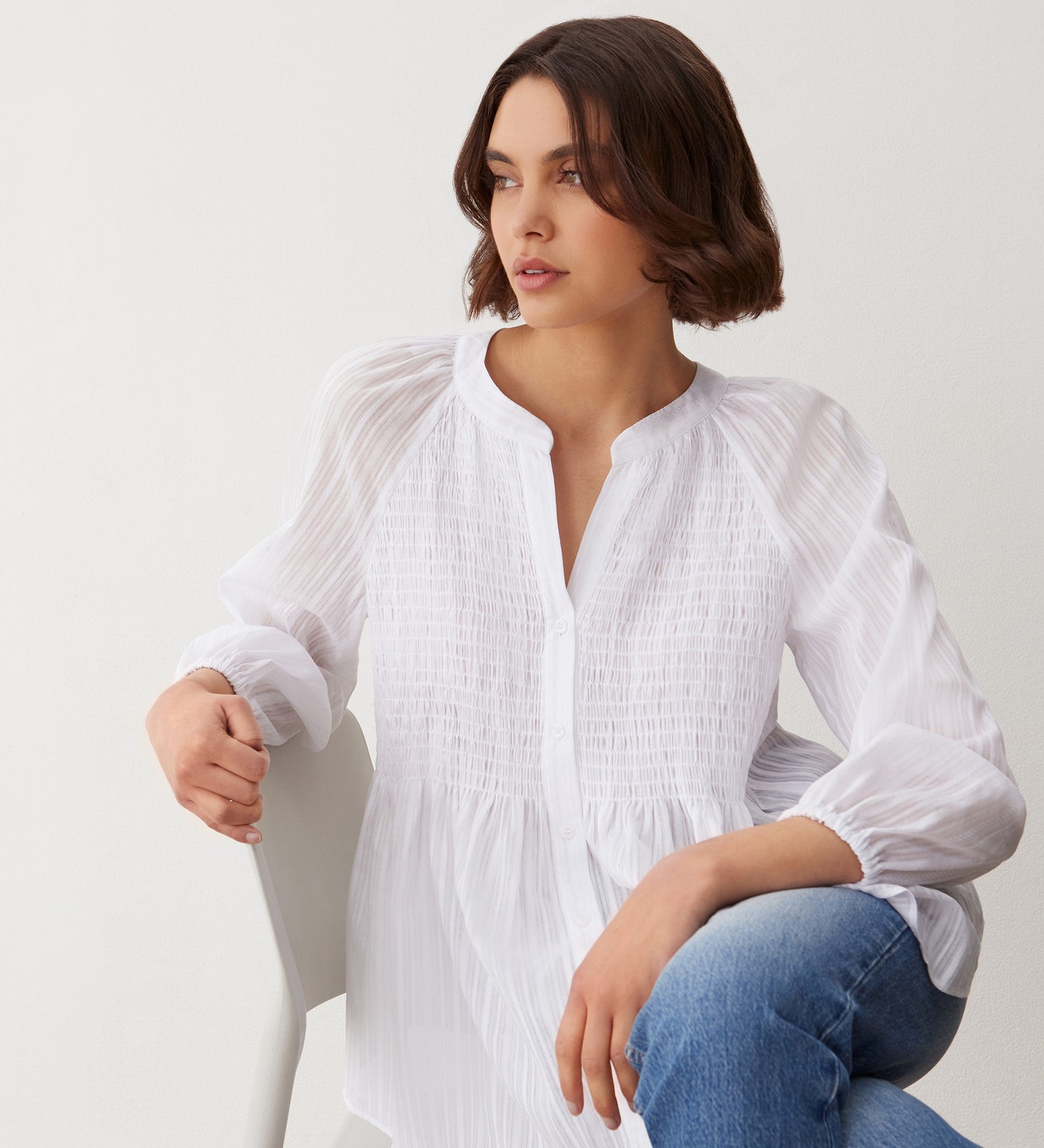 A woman with short brown hair sits on a white chair, wearing the Pixie White Cotton Blouse with a textured finish and blue jeans. She gazes to the side with a relaxed expression against a plain white background.