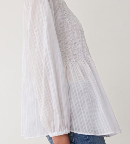 A woman is seen in profile wearing the Pixie White Cotton Blouse—featuring sheer, pleated fabric, shirred bodice, voluminous long sleeves, and a subtle peplum hem—paired with blue jeans against a plain light background.