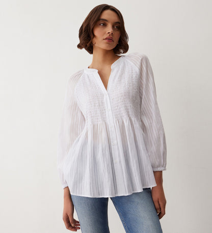 A woman in the Pixie White Cotton Blouse—loose-fitting with long sleeves and vertical stripes—paired with blue jeans stands against a plain light wall, gazing slightly to the side.