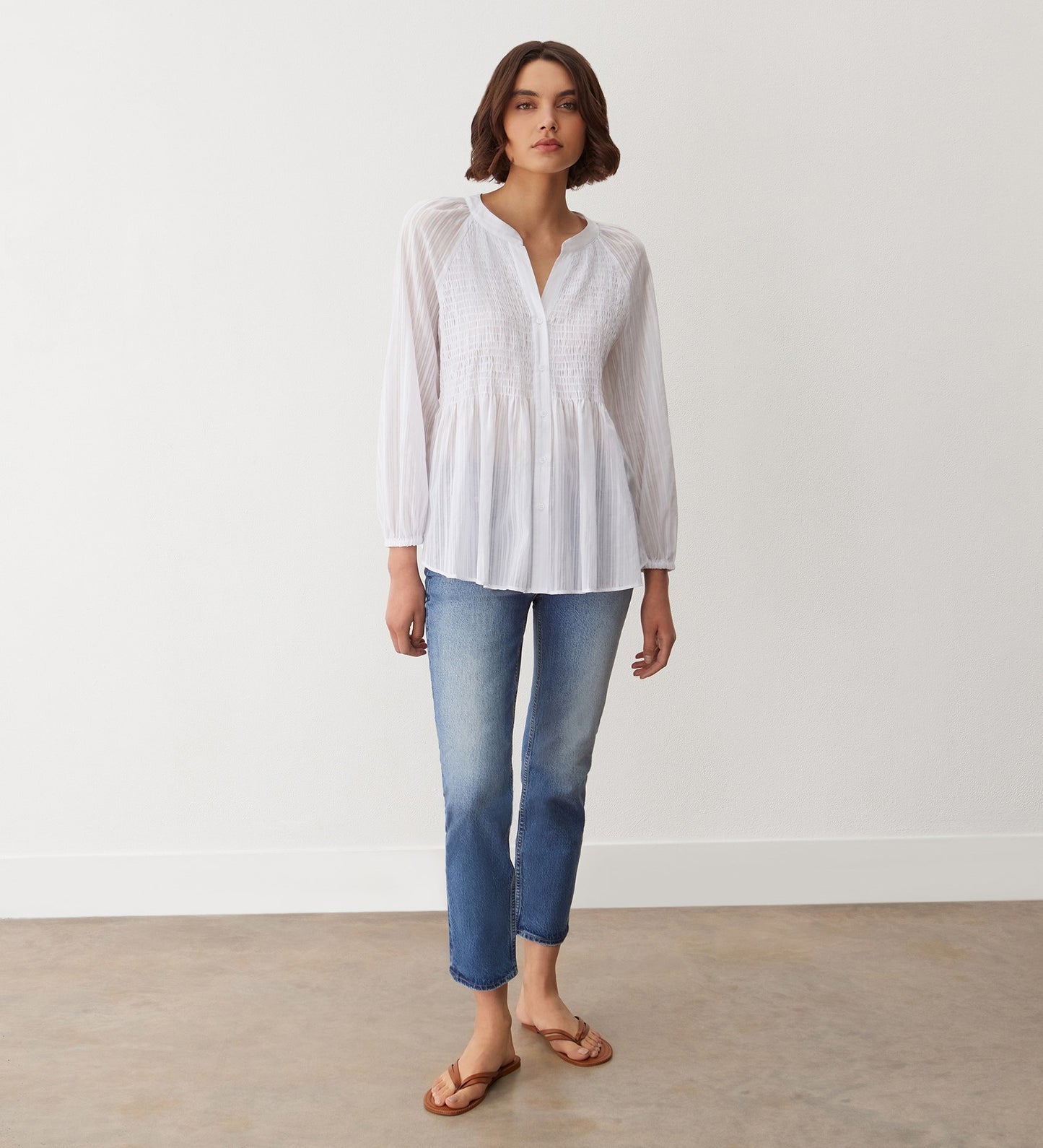 A woman stands indoors against a white wall and concrete floor, wearing the Pixie White Cotton Blouse with pintuck details, styled with blue jeans and tan sandals.