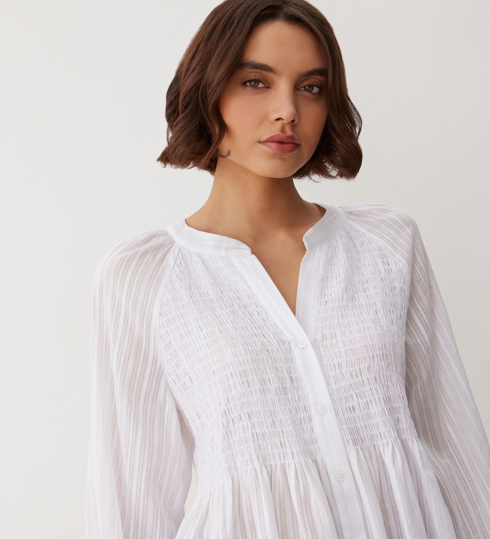 A woman with short brown hair wears the Pixie White Cotton Blouse, a long-sleeve top with pleated details and a shirred bodice, standing against a light background and gazing slightly to the side.