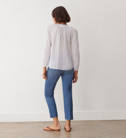 A person stands indoors facing away from the camera, wearing the Pixie White Cotton Blouse, blue jeans, and brown sandals. The background shows a plain white wall and a light-colored floor.