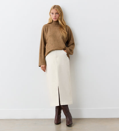 A woman with long blonde hair and a neutral expression stands against a white wall, wearing a loose brown turtleneck sweater, brown boots, and the Rachel Ecru Cotton Rich Denim Midi Skirt featuring a front slit and straight fit.