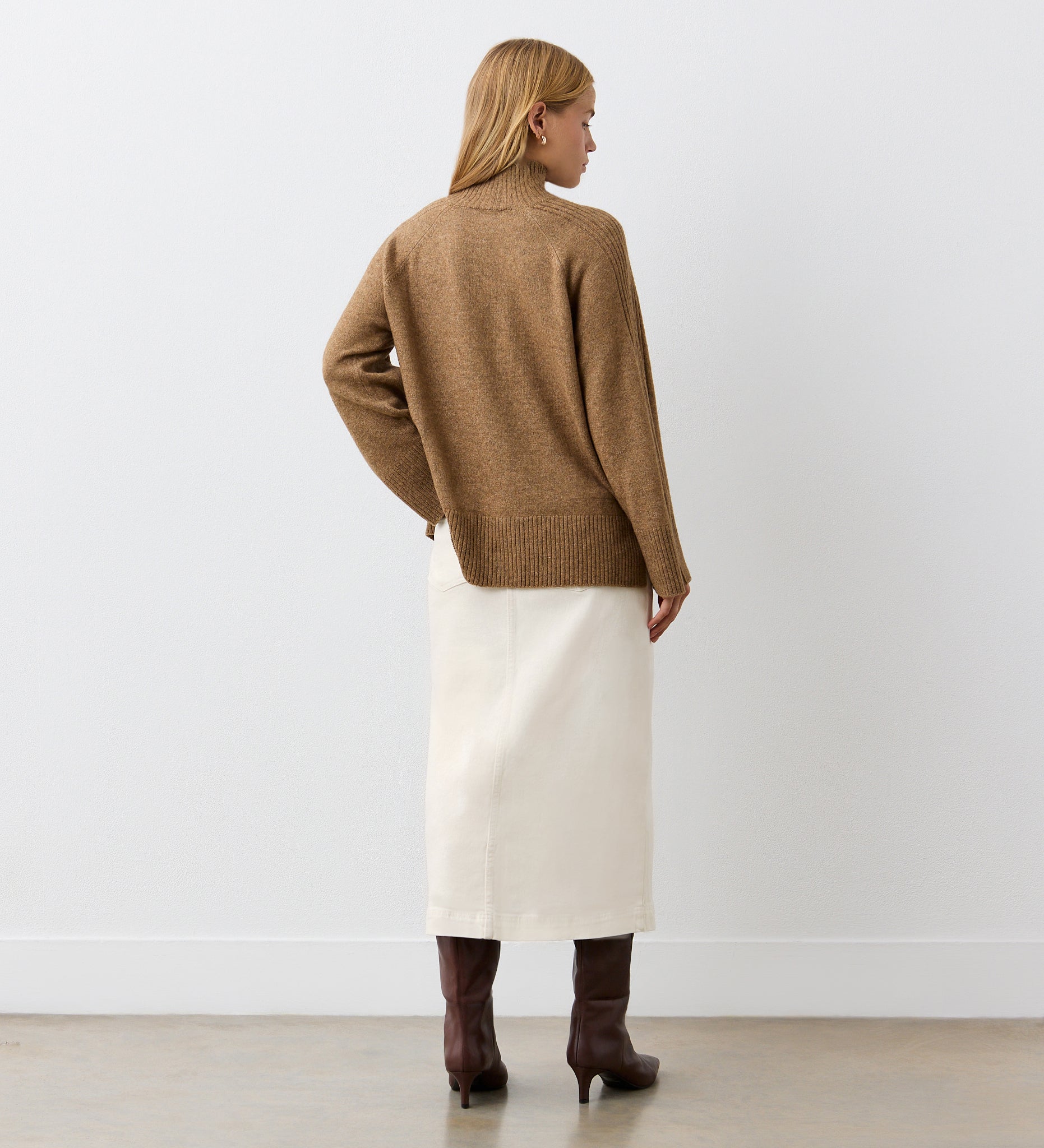 A woman with blonde hair faces away, wearing a brown sweater, brown knee-high boots, and the Rachel Ecru Cotton Rich Denim Midi Skirt in a straight fit, against a plain white wall and light floor.