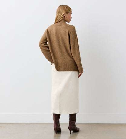 A woman with blonde hair faces away, wearing a brown sweater, brown knee-high boots, and the Rachel Ecru Cotton Rich Denim Midi Skirt in a straight fit, against a plain white wall and light floor.