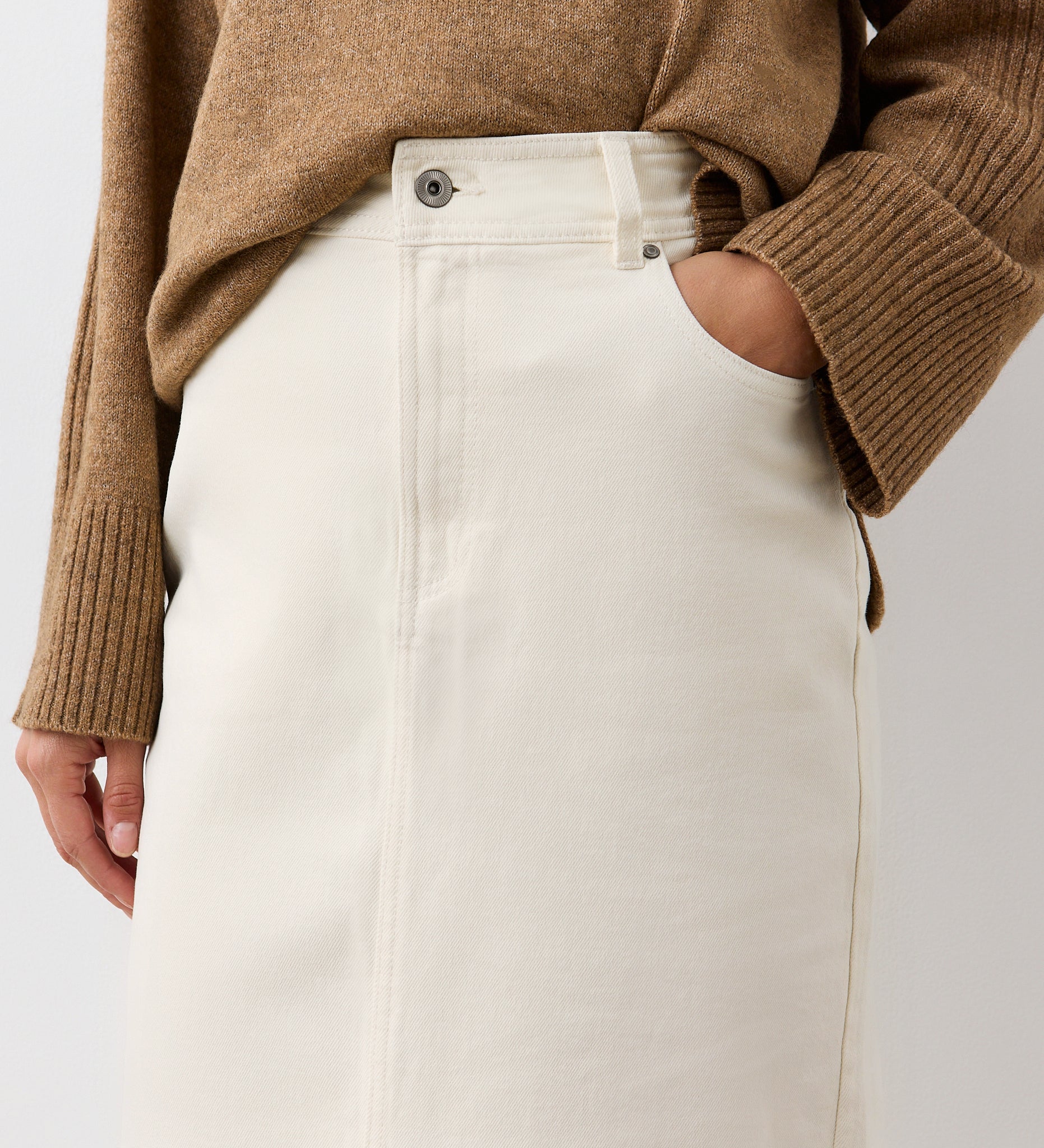 Wearing a brown ribbed sweater with sleeves slightly rolled up and the Rachel Ecru Cotton Rich Denim Midi Skirt, one hand tucked into the pocket of the high-waisted, straight fit cotton-rich denim.