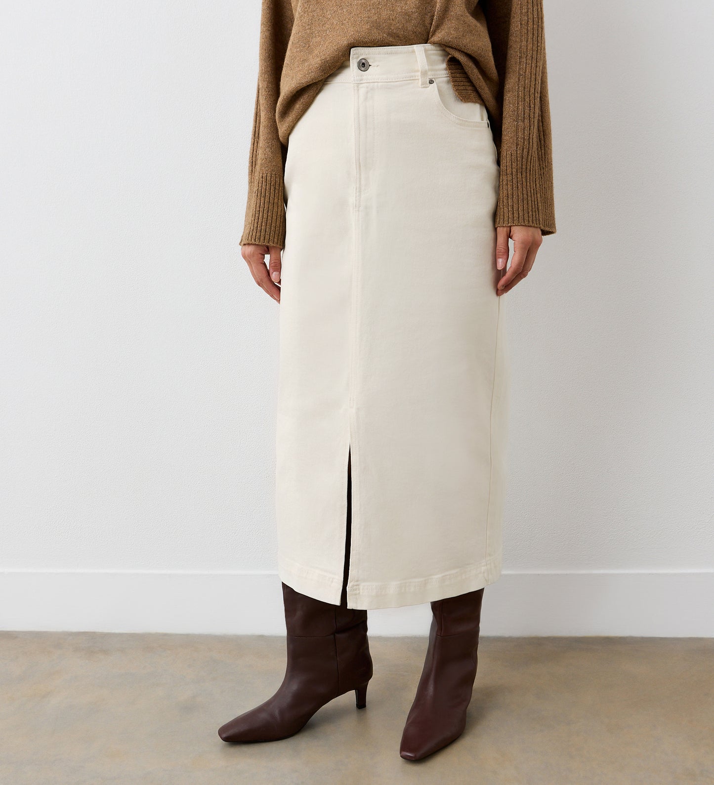 A person wearing a tan sweater, Rachel Ecru Cotton Rich Denim Midi Skirt with a center front slit, and dark brown heeled boots stands against a plain white wall.