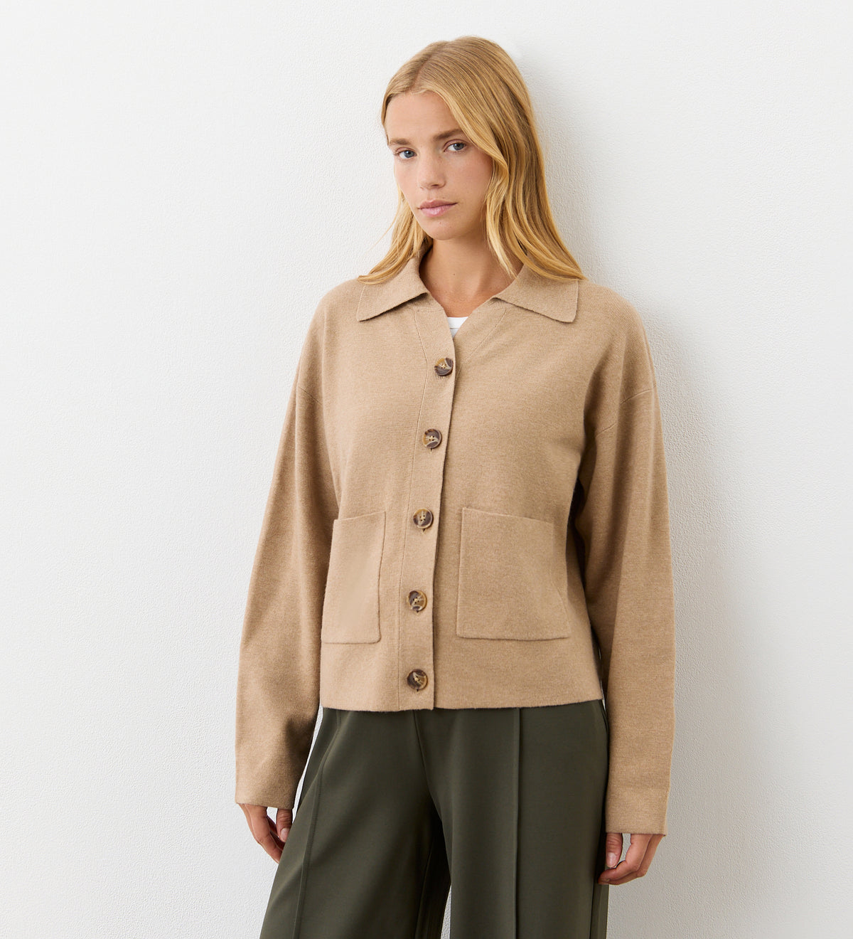 A woman with long blonde hair wears the Raegan Beige Button Front Cardigan, which features functional pockets, paired with dark green trousers as she stands against a plain white wall.