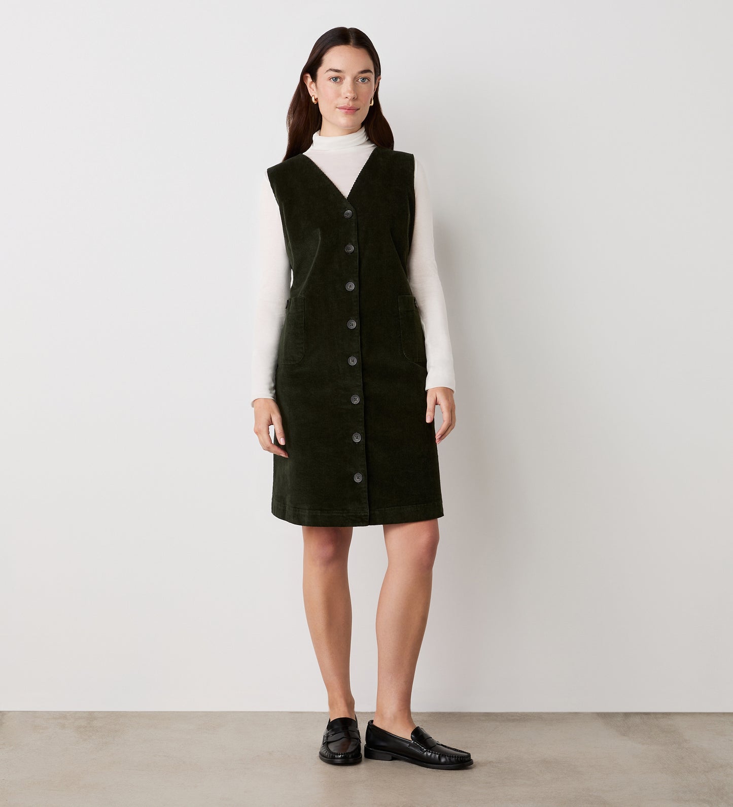 A woman with straight dark hair stands against a plain light wall, wearing the Reneka Khaki Corduroy Knee Length Dress over a white turtleneck and black loafers.