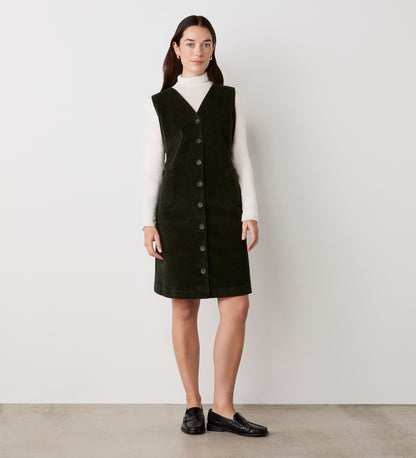 A woman with straight dark hair stands against a plain light wall, wearing the Reneka Khaki Corduroy Knee Length Dress over a white turtleneck and black loafers.