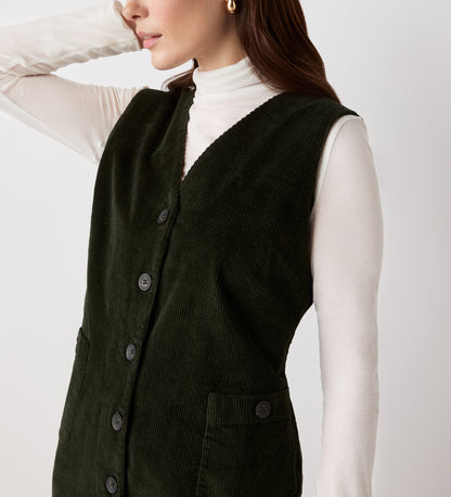 A woman models the Reneka Khaki Corduroy Knee Length Dress over a white turtleneck, standing with one arm bent and touching her head, her face partially out of frame.