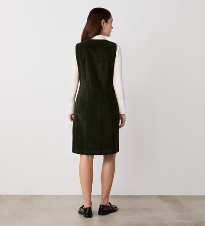 A woman with straight brown hair faces away, wearing the Reneka Khaki Corduroy Knee Length Dress over a white long-sleeve top and black loafers, standing against a plain light background.