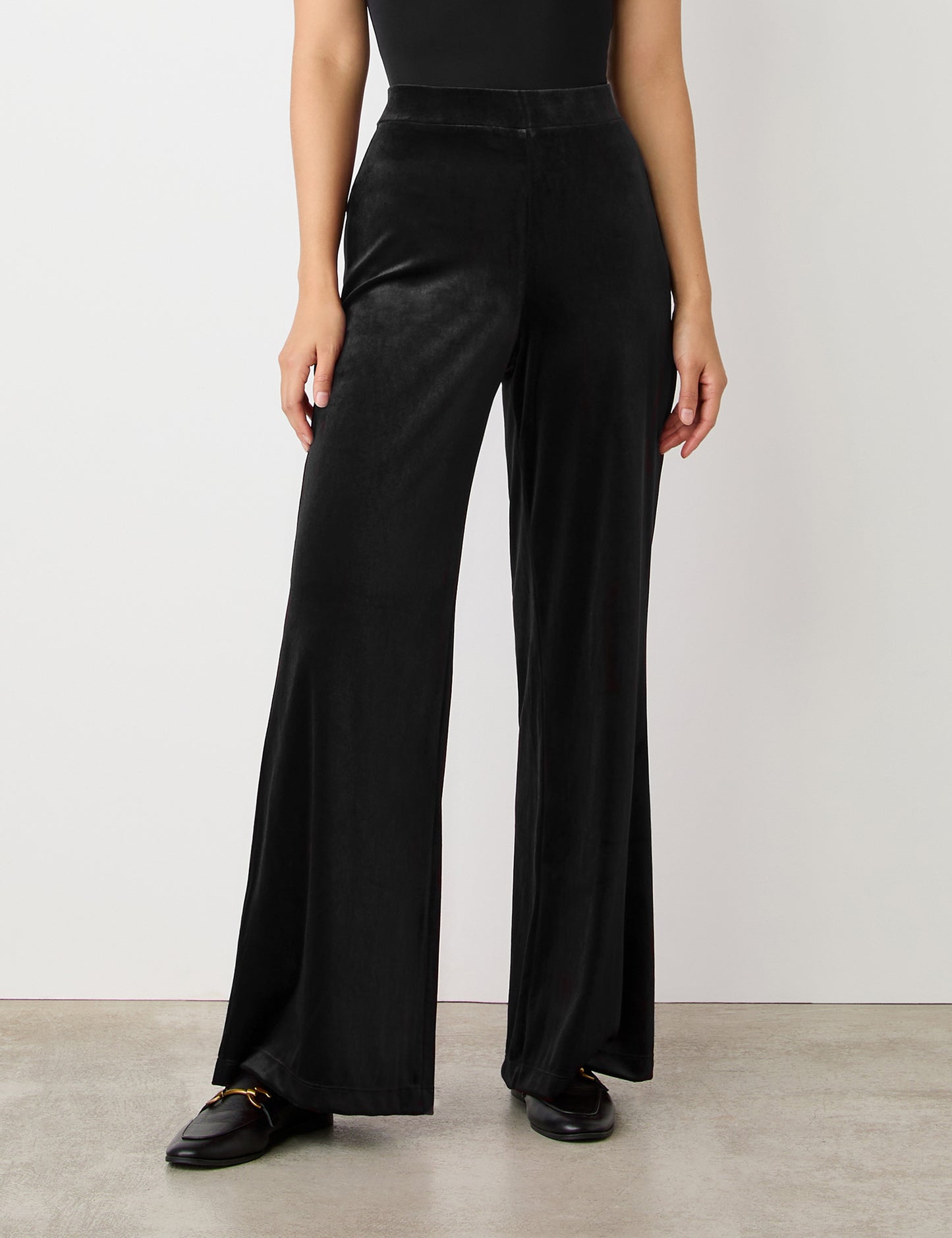 A person models the Reyna Black Velvet Wide Leg Trousers with a high-rise waistband, paired with black loafers, against a white wall. Only their lower torso and legs are visible.