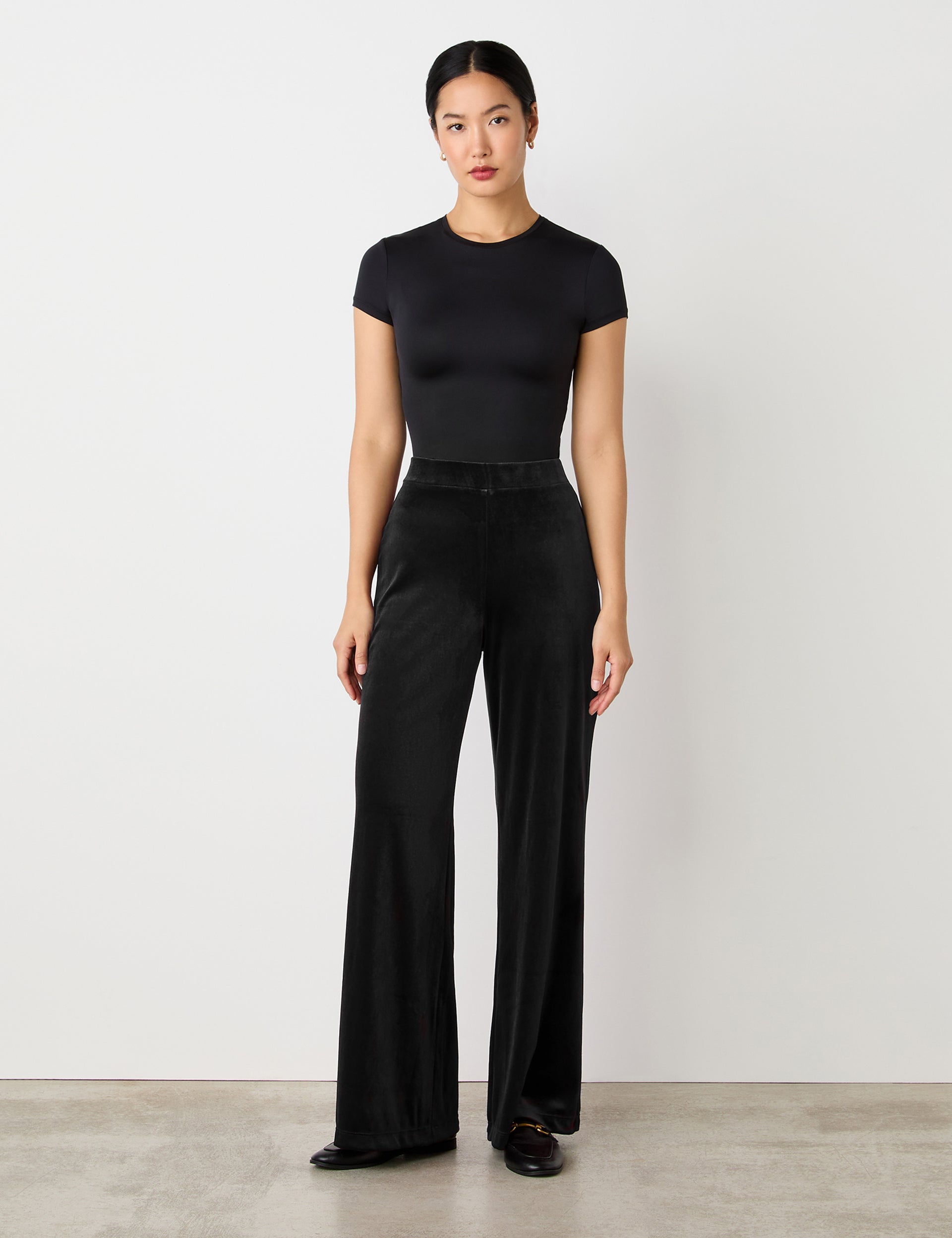 A woman stands against a plain light background, wearing a fitted black top and Reyna Black Velvet Wide Leg Trousers with a high-rise waist and loose silhouette, paired with black shoes.
