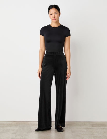 A woman stands against a plain light background, wearing a fitted black top and Reyna Black Velvet Wide Leg Trousers with a high-rise waist and loose silhouette, paired with black shoes.