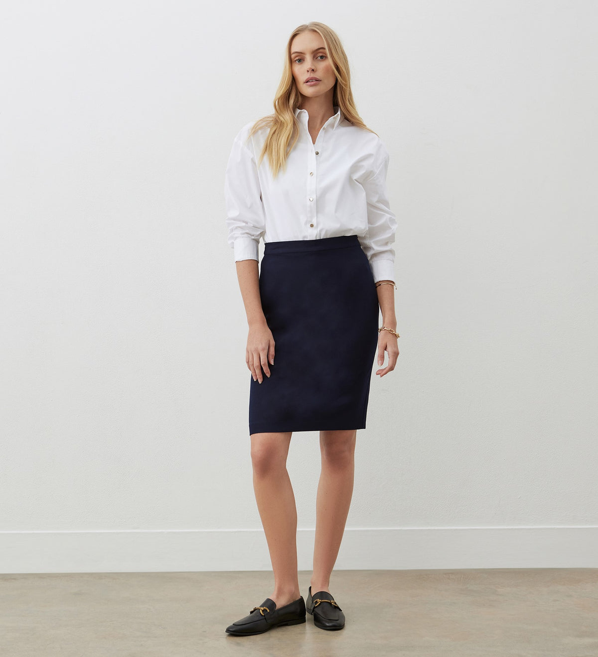 A woman with long blonde hair and a neutral expression stands against a white wall, wearing a white button-up shirt tucked into the Bella Navy Knee Length Skirt and black loafers.