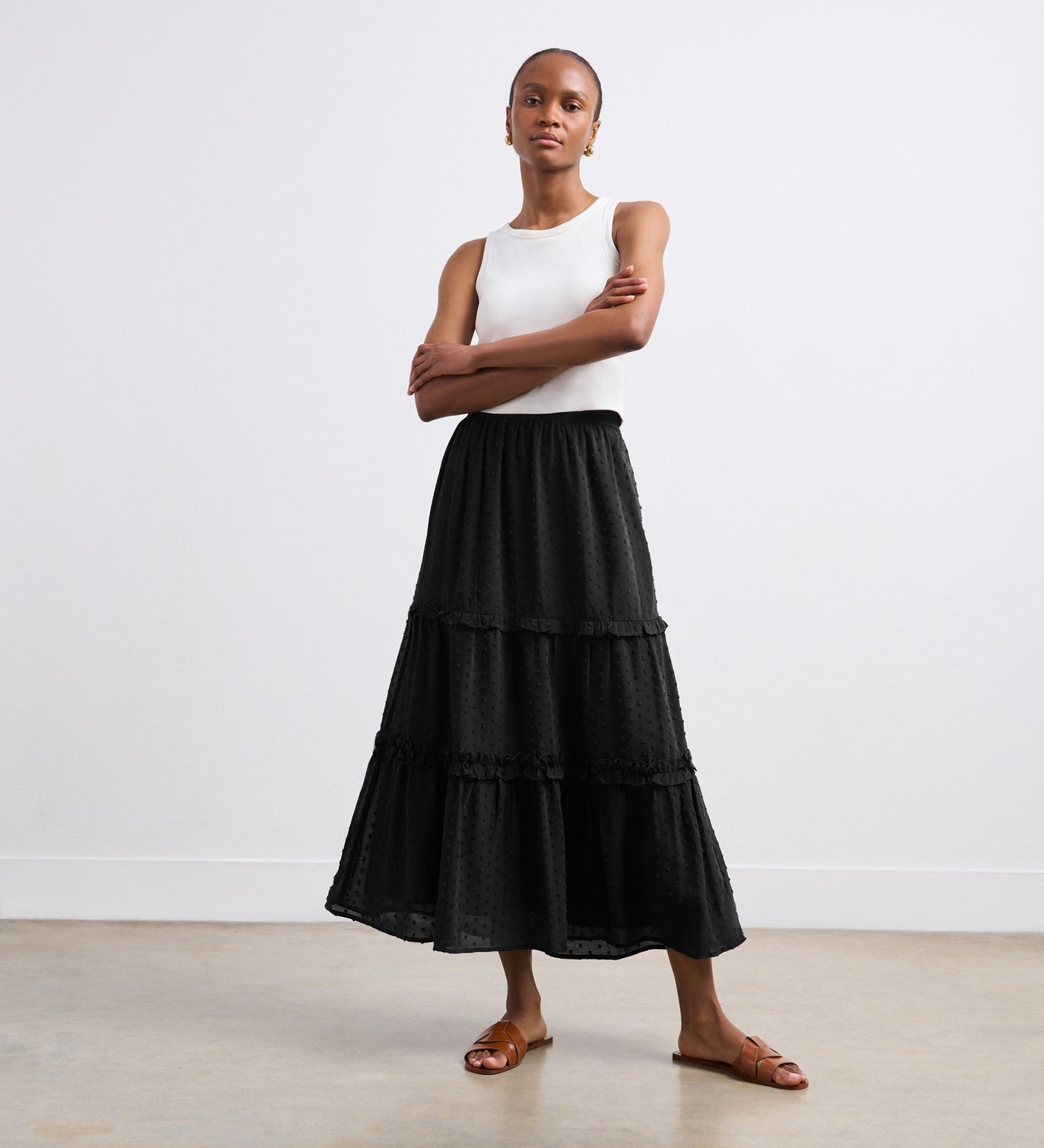 A woman stands with arms crossed, wearing a white sleeveless top, brown sandals, and the Everley Black Tiered Midi Skirt with ruffled detailing. She is posed against a plain white wall and light-colored floor.