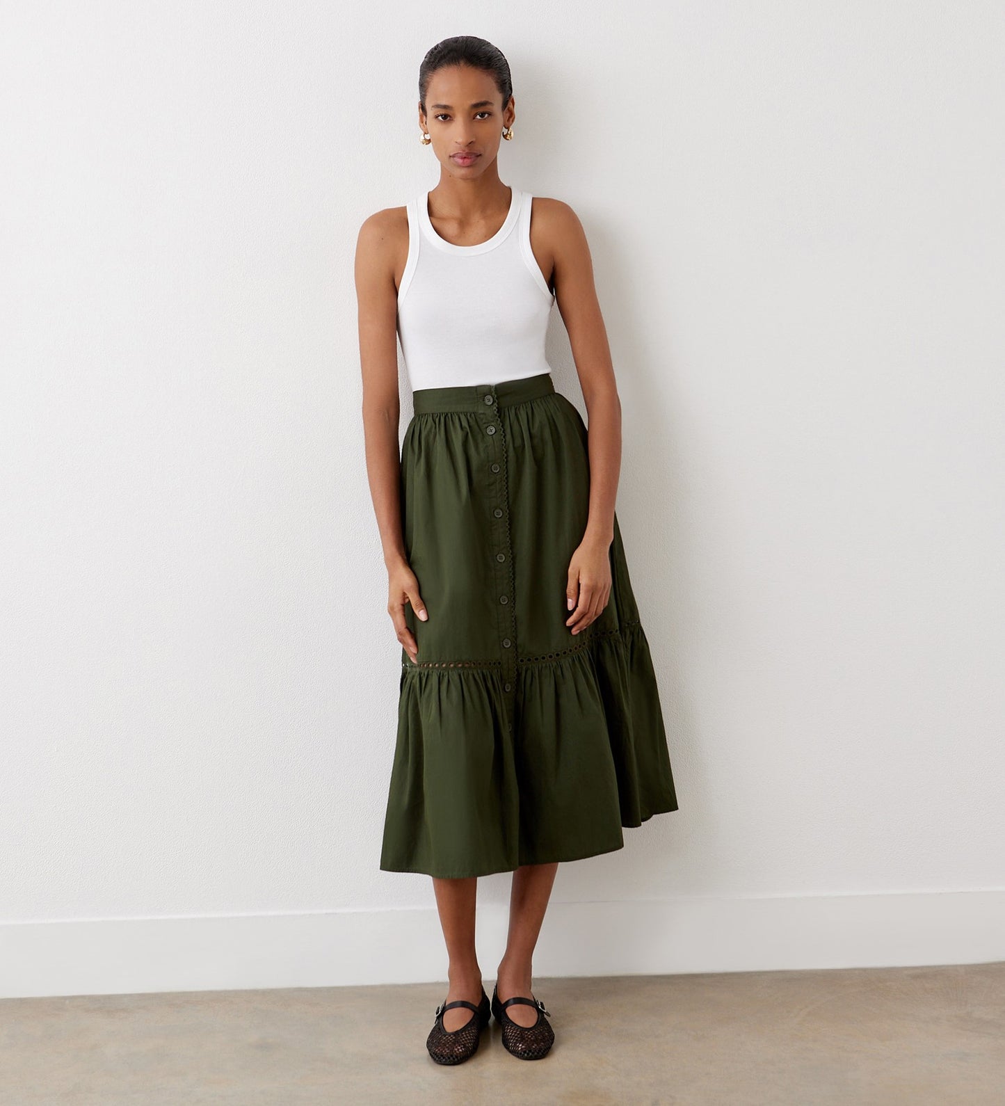A woman stands indoors against a white wall, wearing a white sleeveless tank top, the Maura Khaki Cotton Midi Skirt with a button front, and black shoes. Her hair is pulled back, and her hands rest at her sides.