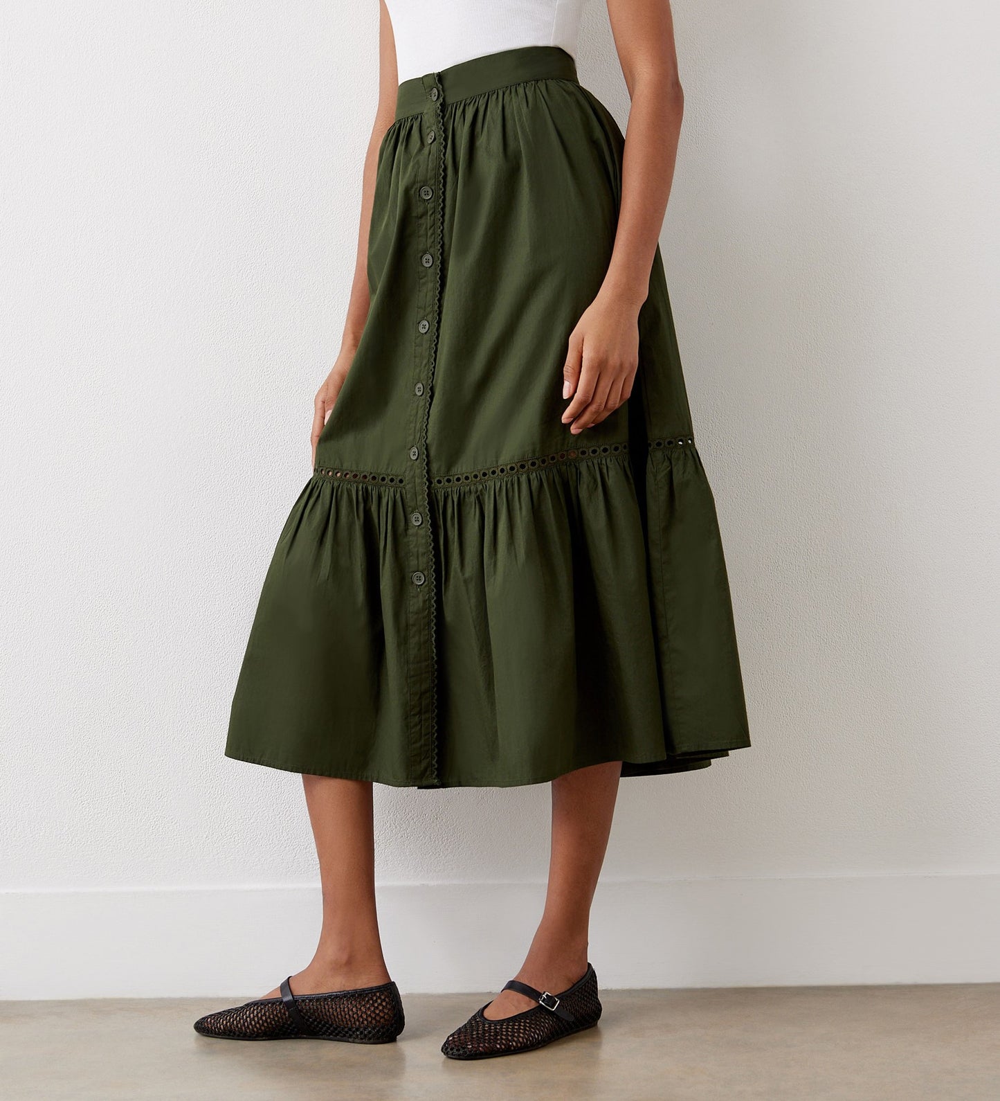 A woman stands against a white wall wearing a sleeveless white top, the Maura Khaki Cotton Midi Skirt featuring gathered ruffles and a tiered hemline, and black mesh ballet flats.