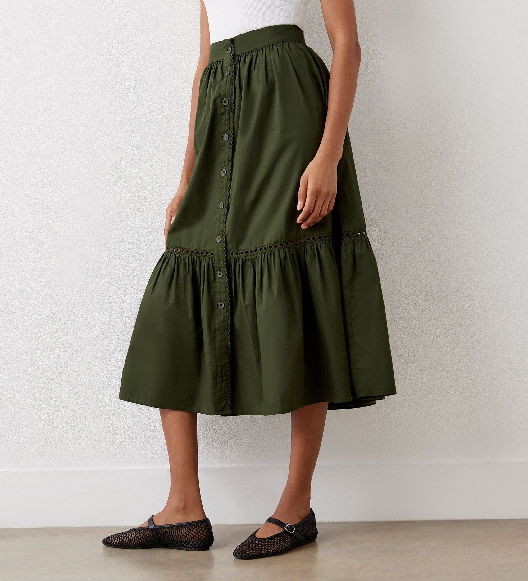 A woman stands against a white wall wearing a sleeveless white top, the Maura Khaki Cotton Midi Skirt featuring gathered ruffles and a tiered hemline, and black mesh ballet flats.