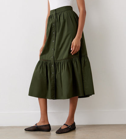 A woman stands against a white wall wearing a sleeveless white top, the Maura Khaki Cotton Midi Skirt featuring gathered ruffles and a tiered hemline, and black mesh ballet flats.