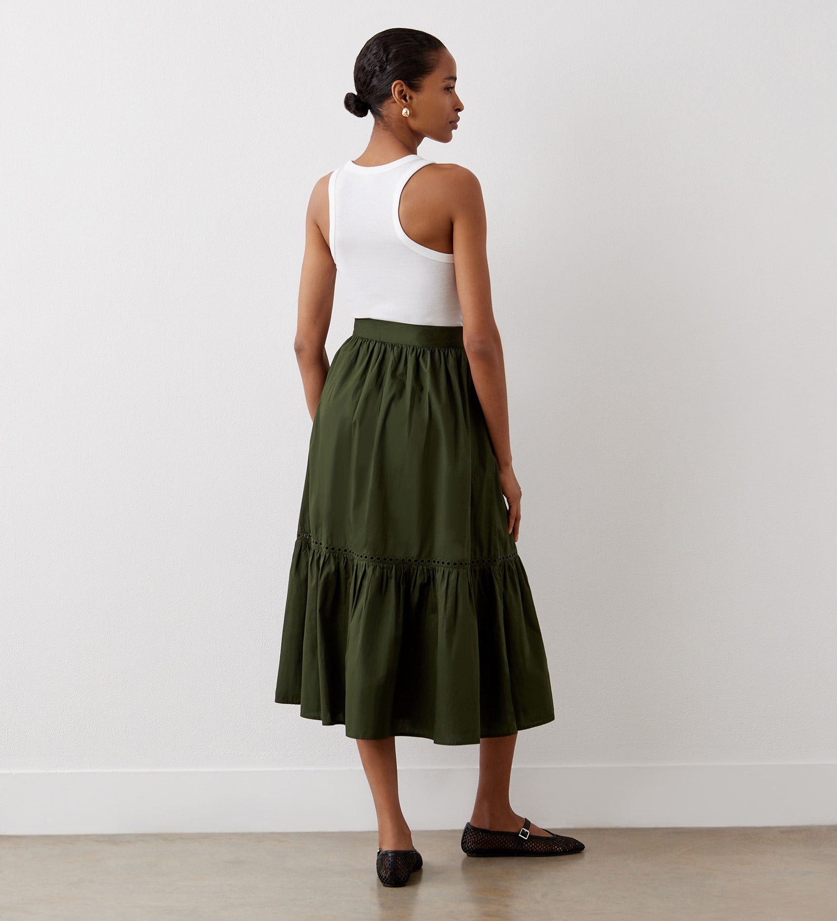 A woman stands with her back to the camera, wearing a white sleeveless top and the Maura Khaki Cotton Midi Skirt with a ruffled tiered hemline, paired with black flats. She poses against a plain white wall on a light floor.