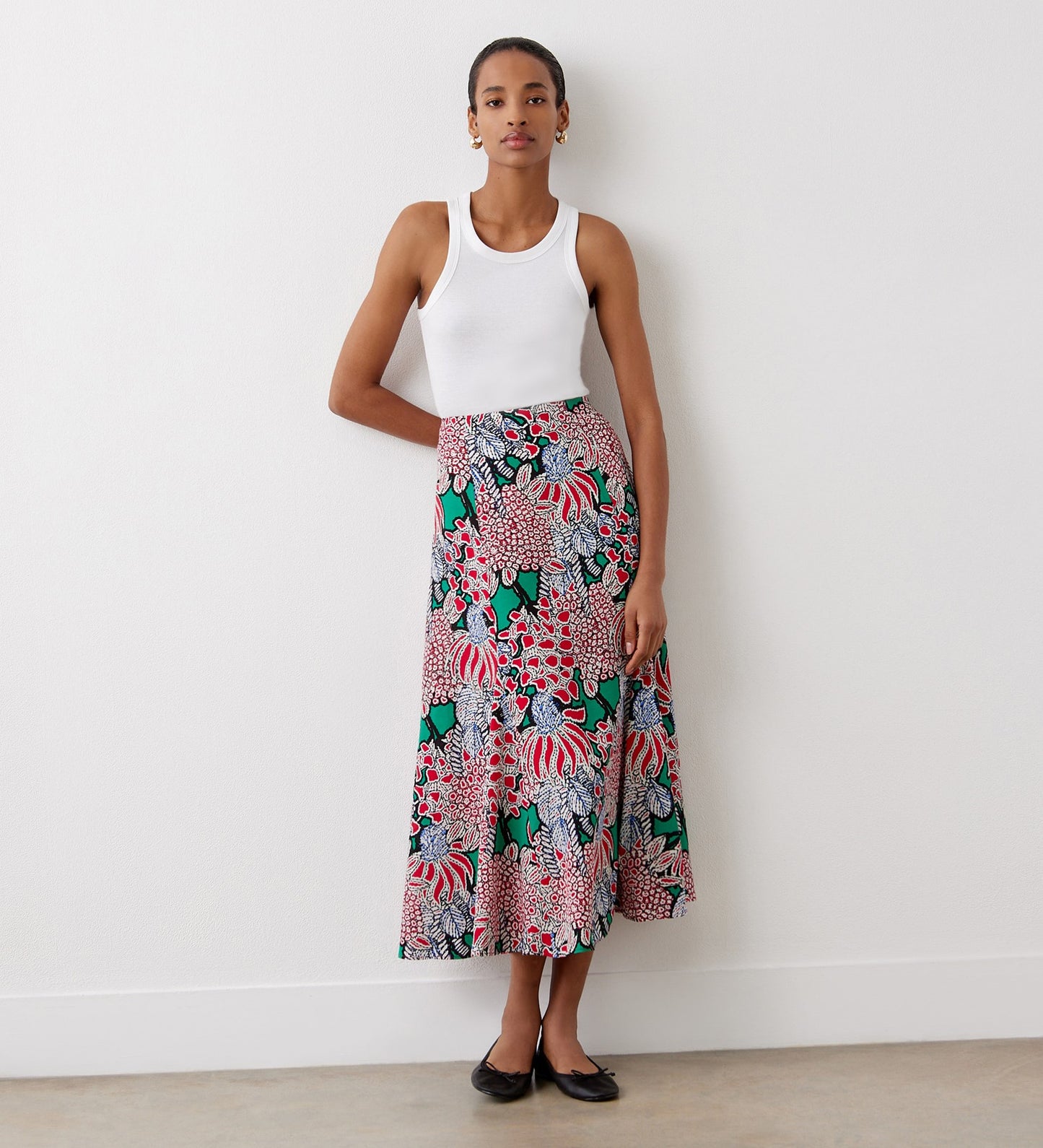 A woman stands against a white wall wearing a white sleeveless tank top, the Jessie Pink Bloom Midi Skirt with red, pink, green, and black floral patterns, and black ballet flats.
