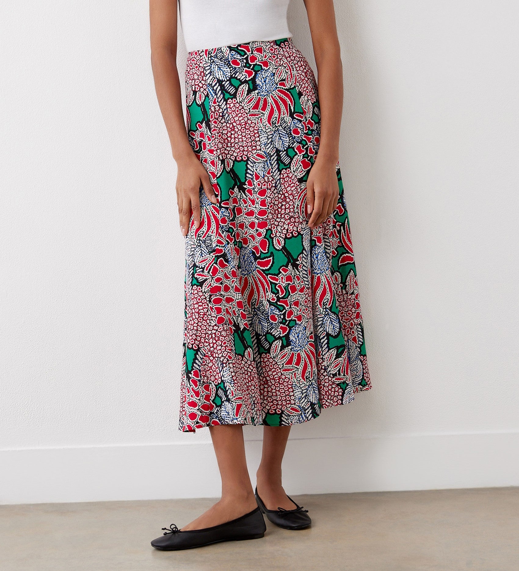 A person stands against a white wall in a white sleeveless top, black ballet flats, and the Jessie Pink Bloom Midi Skirt featuring a vibrant floral pattern in red, green, black, and white.