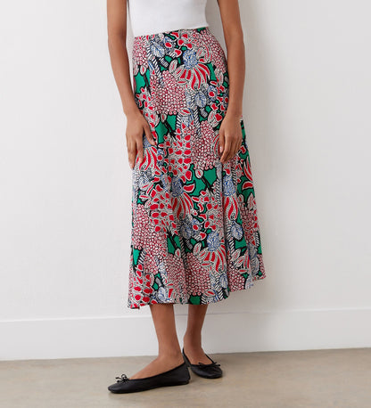 A person stands against a white wall in a white sleeveless top, black ballet flats, and the Jessie Pink Bloom Midi Skirt featuring a vibrant floral pattern in red, green, black, and white.