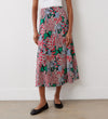 A person wearing a white top and the Jessie Pink Bloom Midi Skirt featuring vibrant floral prints, paired with black ballet flats, stands against a plain white wall.