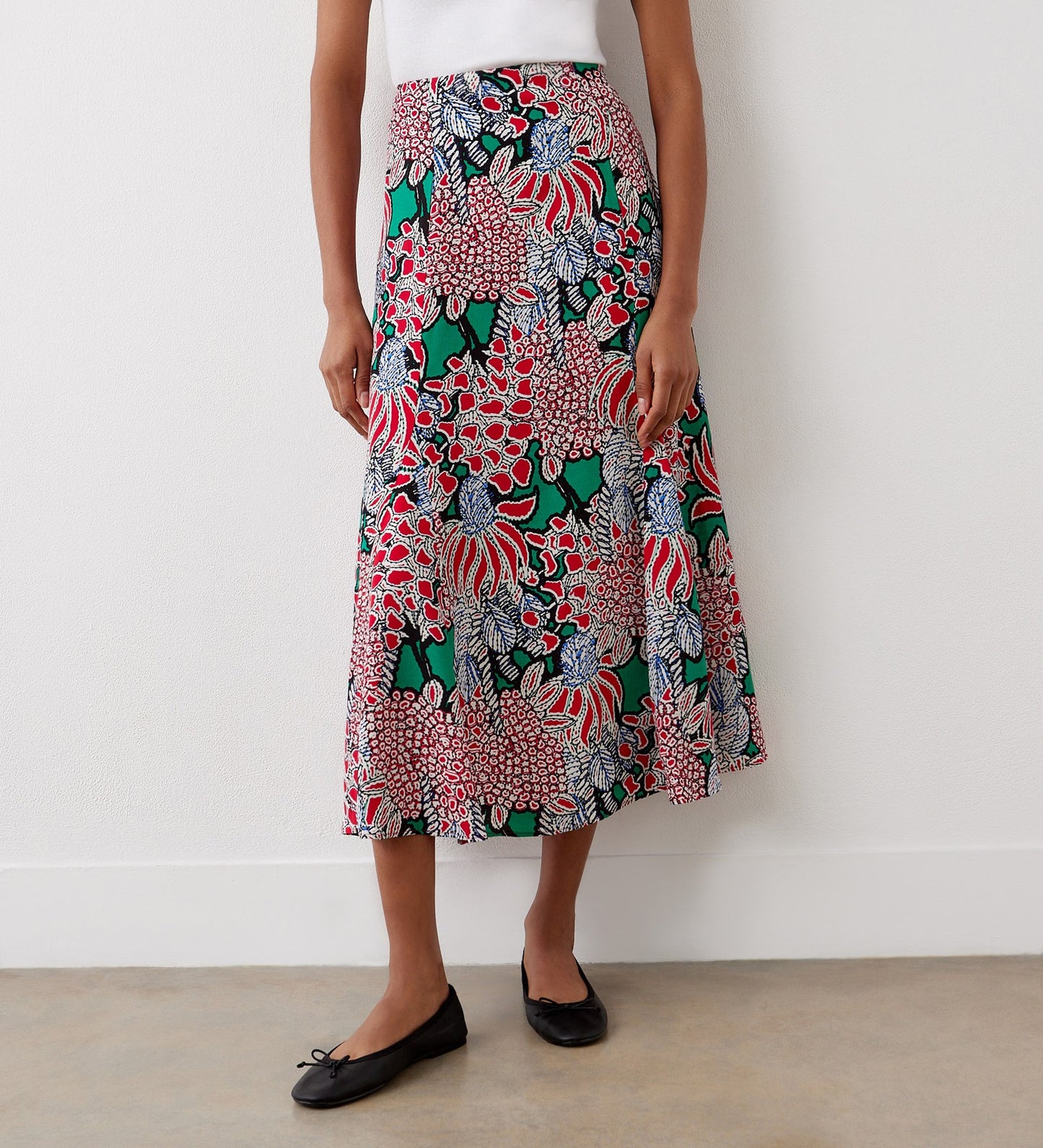 A person wearing a white top and the Jessie Pink Bloom Midi Skirt featuring vibrant floral prints, paired with black ballet flats, stands against a plain white wall.
