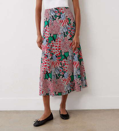 A person wearing a white top and the Jessie Pink Bloom Midi Skirt featuring vibrant floral prints, paired with black ballet flats, stands against a plain white wall.