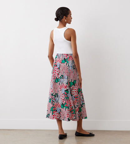 A woman stands facing a white wall, wearing a white sleeveless top, black flats, and the Jessie Pink Bloom Midi Skirt—a colorful, bias-cut floral skirt. Her hair is tied back and she is on a light-colored floor.