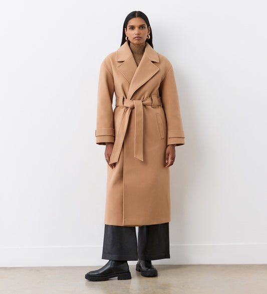 A woman with straight dark hair and a neutral expression stands against a white wall wearing the Sally Camel Long Coat over a brown turtleneck, black wide-leg pants, and chunky boots.