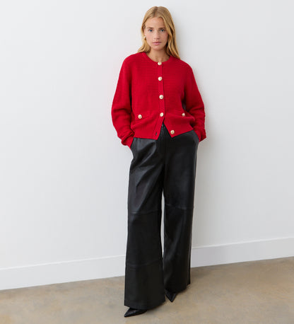 A woman with long blonde hair stands against a white wall, hands in pockets, wearing the Saskia Red Button Up Cotton Cardigan with gold crest buttons, high-waisted black wide-leg leather pants, and pointed black shoes.
