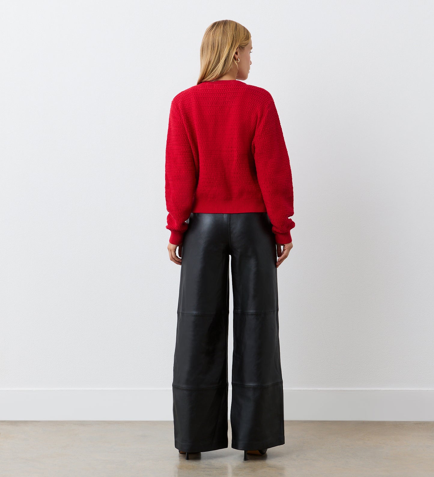 A person with blonde hair, wearing the Saskia Red Button Up Cotton Cardigan and wide-leg black leather pants, stands facing a white wall with their back to the camera.