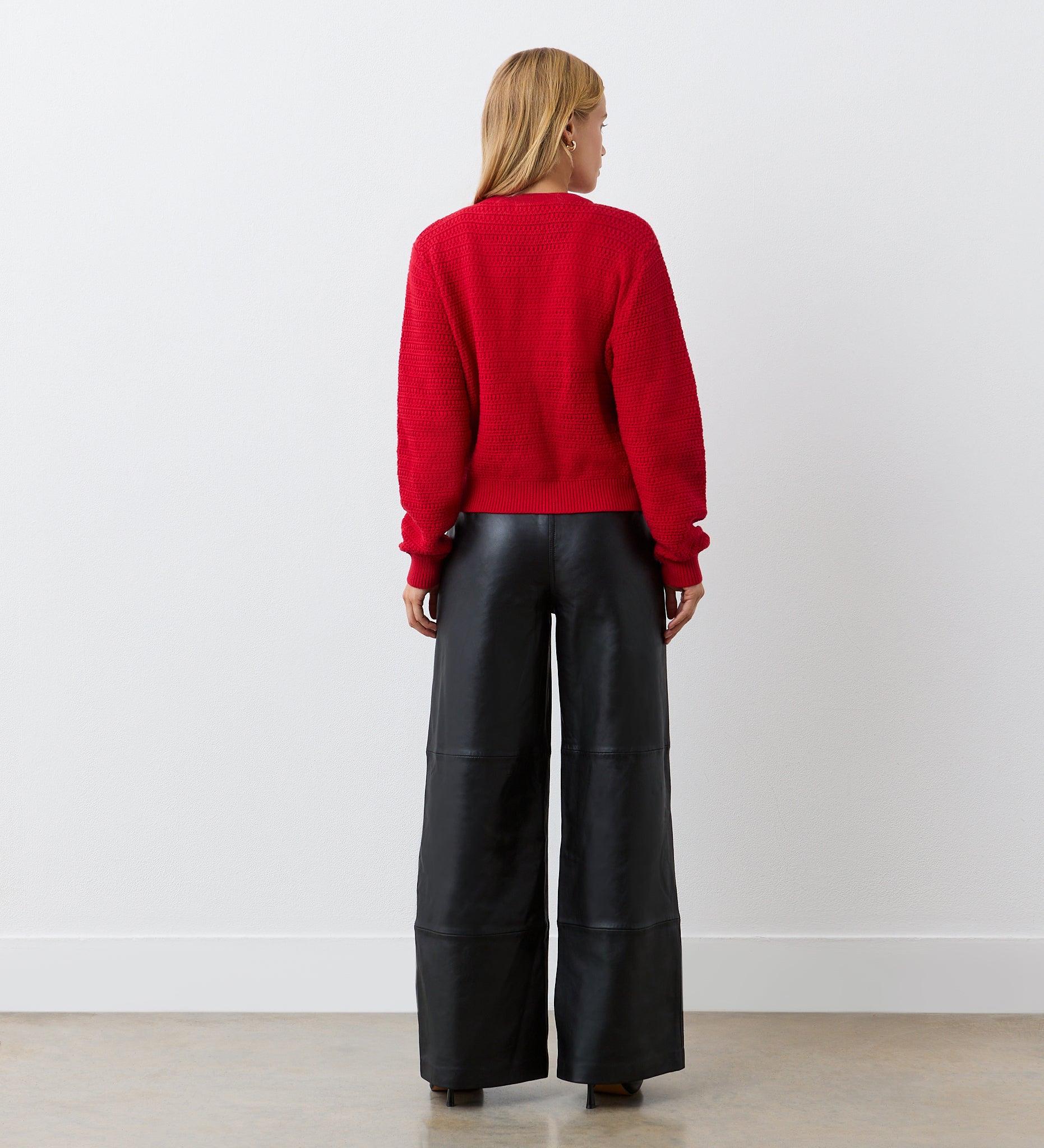 A person with blonde hair, wearing the Saskia Red Button Up Cotton Cardigan and wide-leg black leather pants, stands facing a white wall with their back to the camera.