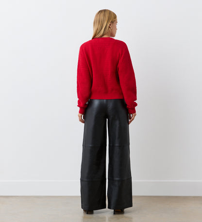 A person with blonde hair, wearing the Saskia Red Button Up Cotton Cardigan and wide-leg black leather pants, stands facing a white wall with their back to the camera.