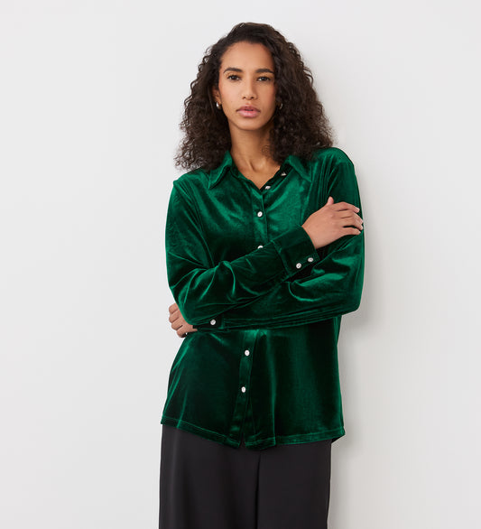 A woman with curly hair wears the Sayge Bottle Green Velvet Shirt with silver buttons and black pants, standing against a plain white background with her arms lightly folded.