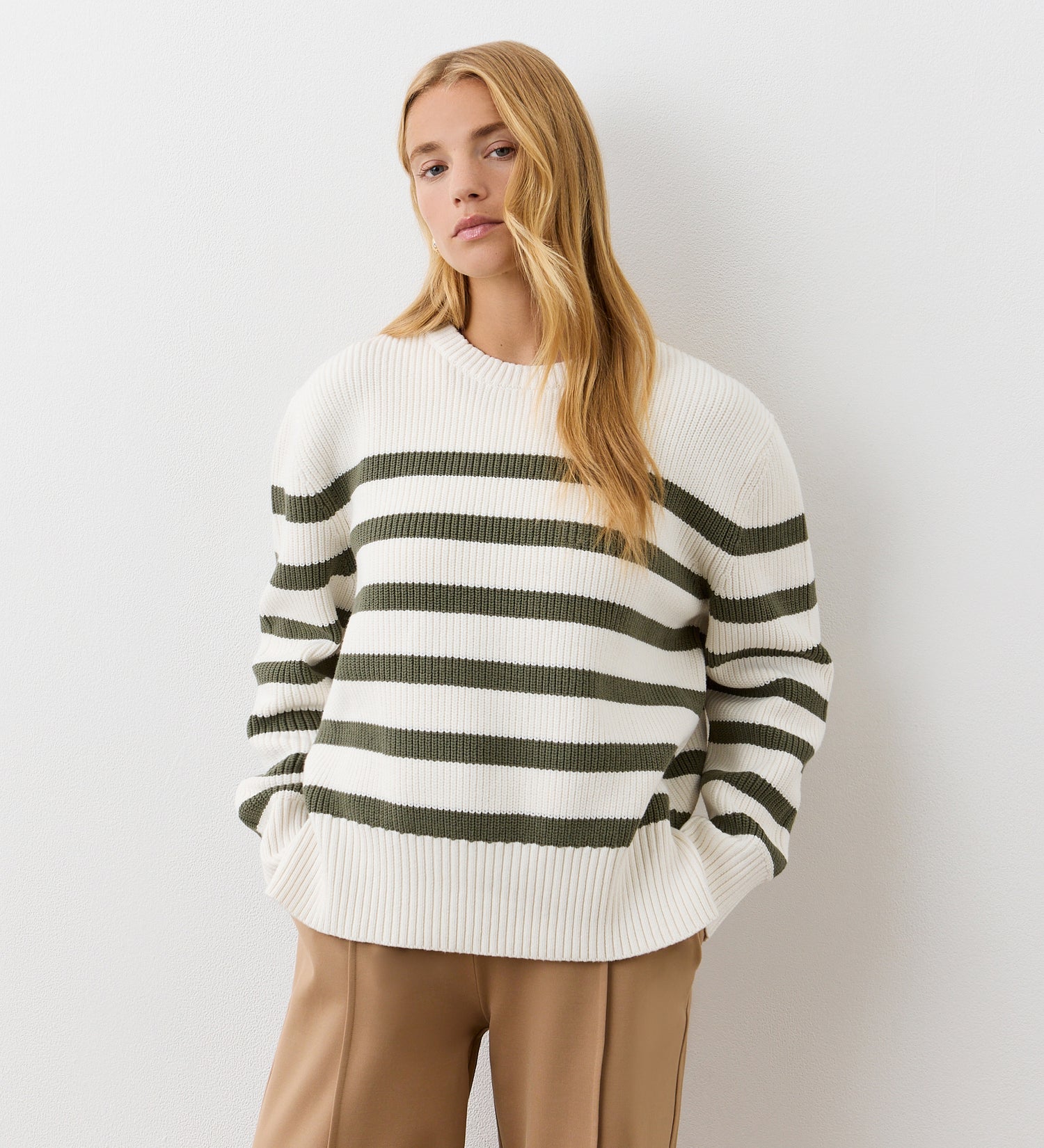 A woman with long blonde hair stands against a plain white wall, hands in her pockets, wearing tan pants and the Serena Khaki Stripe Knitted Sweater, looking calmly at the camera.