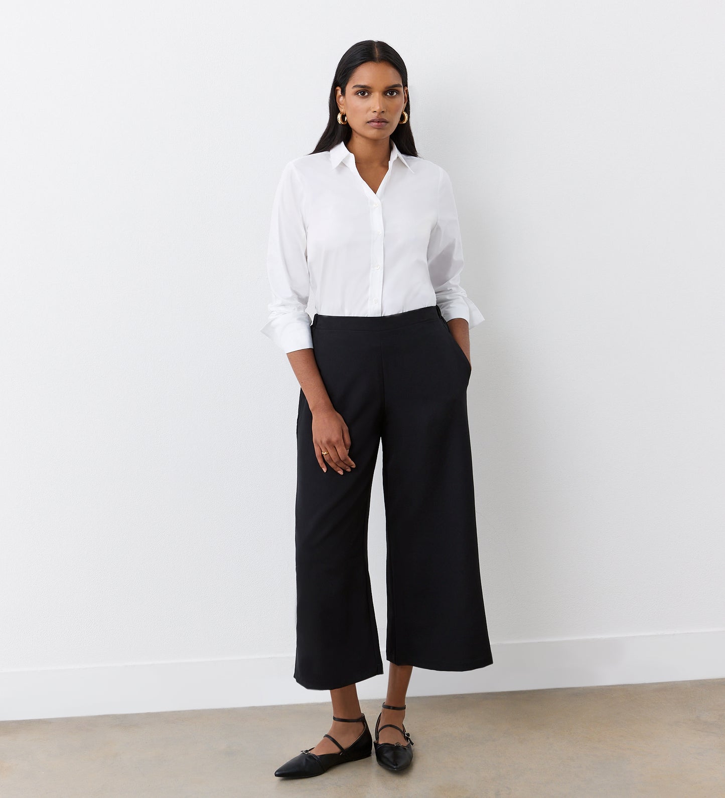A woman stands against a white wall wearing a white button-up shirt and Simone Black Cropped Wide Leg Trousers with an elastic waistband. She pairs them with black pointed flats, gold hoop earrings, and has her hands in her pockets.