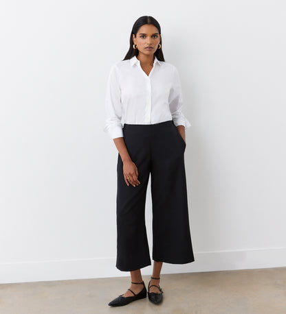 A woman stands against a white wall wearing a white button-up shirt and Simone Black Cropped Wide Leg Trousers with an elastic waistband. She pairs them with black pointed flats, gold hoop earrings, and has her hands in her pockets.