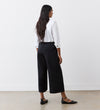 A woman stands indoors facing away, wearing a white long-sleeve blouse and the Simone Black Cropped Wide Leg Trousers with an elasticated waistband, paired with black ankle strap flats on a light floor against a plain white wall.