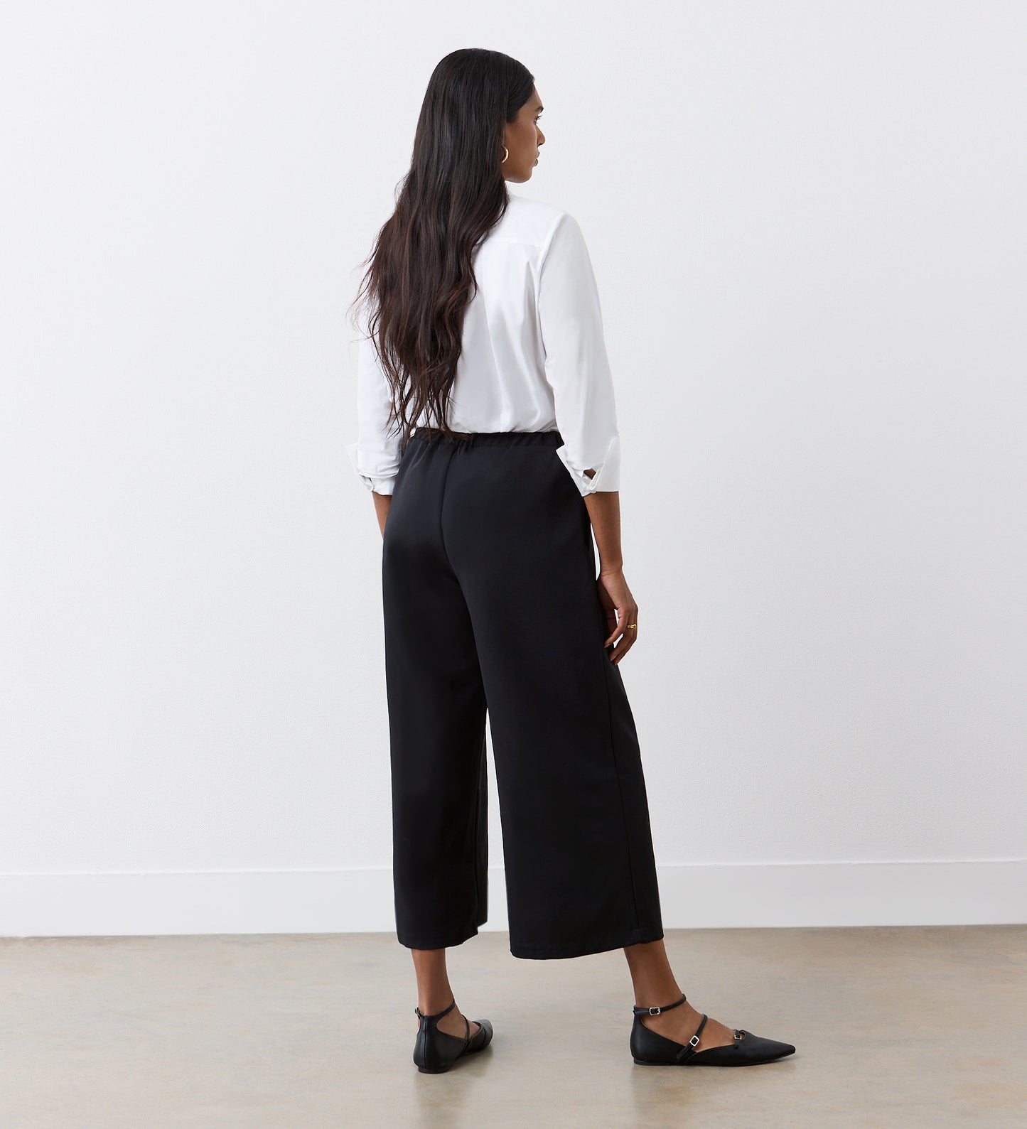A woman stands indoors facing away, wearing a white long-sleeve blouse and the Simone Black Cropped Wide Leg Trousers with an elasticated waistband, paired with black ankle strap flats on a light floor against a plain white wall.