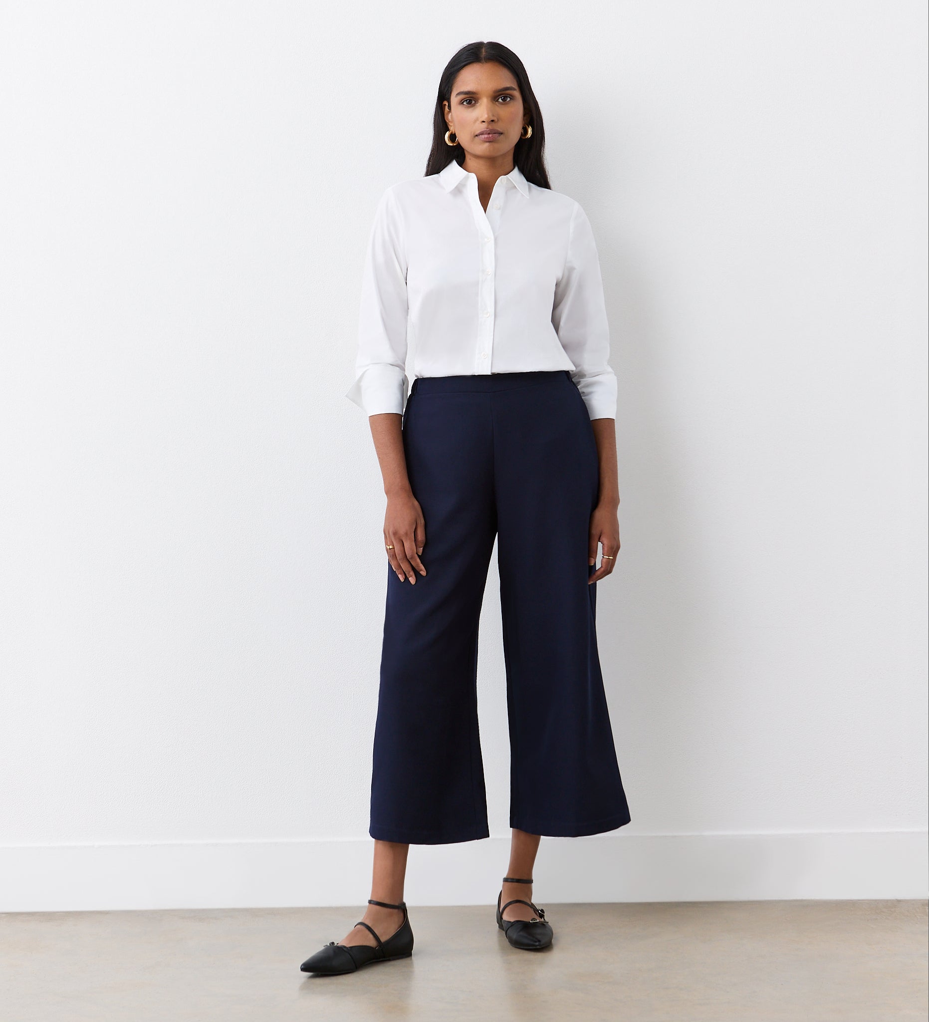 A woman with long dark hair and a neutral expression stands against a white wall wearing Simone Navy Cropped Wide Leg Trousers, a white button-down shirt, black flats, and gold hoop earrings—an effortlessly stylish wardrobe essential.