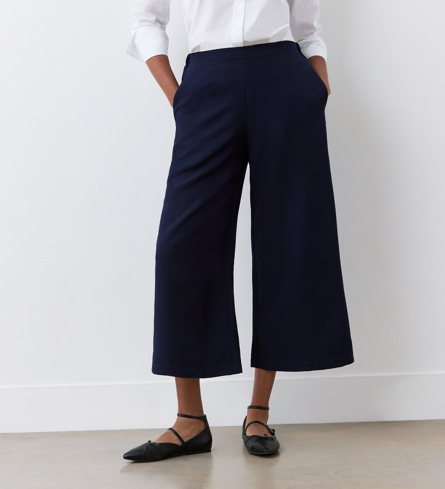 A person stands against a white wall with hands in their pockets, wearing a white long-sleeve blouse, black flats, and the Simone Navy Cropped Wide Leg Trousers featuring an elasticated waistband.